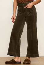 Load image into Gallery viewer, A woman wearing dark olive brown wide-leg corduroy pants with a high waist, front patch pockets, and cuffed hems, paired with brown suede mule slide sandals.