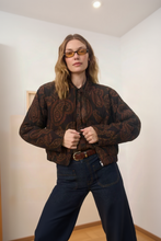 Load image into Gallery viewer, A woman wearing a brown and black paisley patterned bomber jacket, dark blue jeans, and amber-tinted glasses, posing against a neutral indoor background.