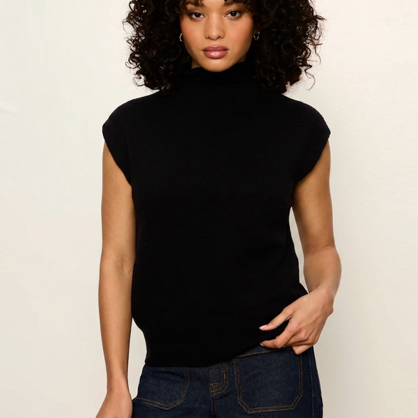 A woman wearing a black sleeveless mock neck knit top and dark blue jeans, standing against a plain off-white wall.