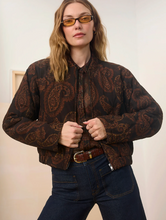 Load image into Gallery viewer, A woman wearing a brown and black paisley patterned bomber jacket, dark blue jeans, and amber-tinted glasses, posing against a neutral indoor background.
