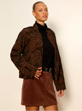 Load image into Gallery viewer, A woman modeling a brown paisley patterned jacket over a black turtleneck and a brown mini skirt, standing against a plain off-white background.
